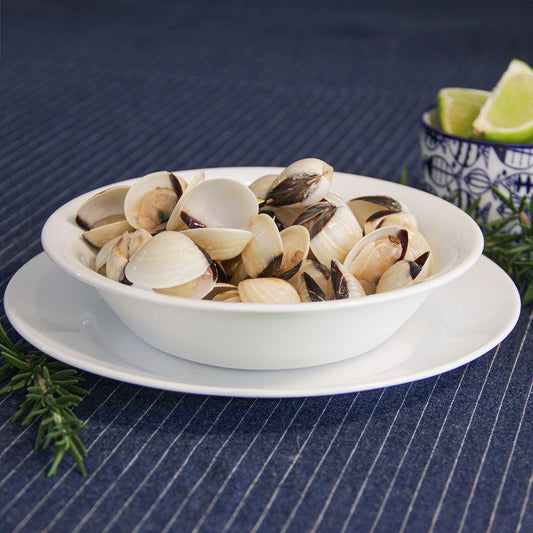 Un plato de almejas frescas servido en un bol blanco, con rodajas de lima y ramitas de romero al lado, sobre un mantel de rayas azules. Ideal para recetas de mariscos con almejas.