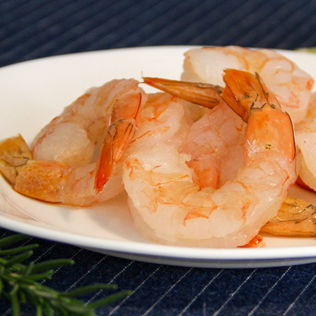 Primer plano de camarones pink pelados y cocidos en un plato blanco, con hierbas aromáticas y una guarnición de lima, presentados sobre un mantel de tela azul oscuro con rayas. Ideal para destacar platos de mariscos con camarón pink en menús y recetas.