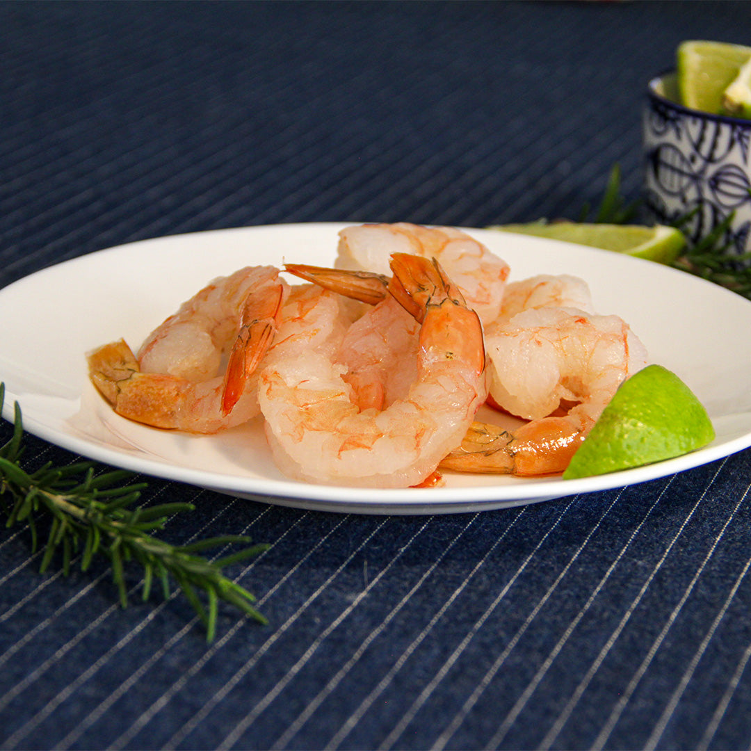 Un plato con camarones pink cocidos, acompañados de rodajas de lima y romero fresco sobre un mantel azul a rayas, perfecto para menús de mariscos.