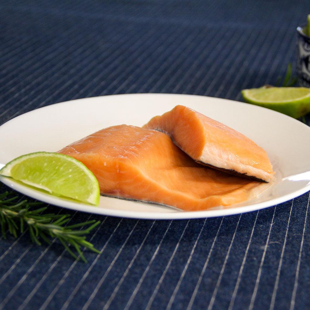 Dos trozos de salmón fresco en un plato blanco, con una rodaja de lima al lado y romero fresco, presentados sobre un mantel azul con rayas. Perfecto para artículos o menús que resalten recetas saludables con salmón.